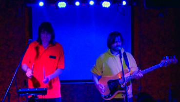 Preview: Bandfoto sunflowerstarfish: Zwei Musiker bei Live-Konzert  Zwei Musiker stehen bei einem Live-Auftritt auf einer blau beleuchteten Bühne. Links spielt ein Musiker an einem elektronischen Instrument, rechts singt ein Musiker am Mikrofon und spielt E-Bass.
