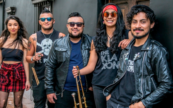 Foto der Band The Klaxon. Fünf Bandmitglieder posieren lässig vor einer schwarzen Wand. Zwei Männer in Lederjacken und Sonnenbrillen halten Trompeten. Eine Frau mit langen dunklen Haaren trägt ein rotes Stirnband und ein T-Shirt mit dem Bandnamen "The Kla