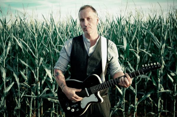 Bandfoto Wayne Jackson: Ein Mann steht in einem hohen Maisfeld und spielt eine E-Gitarre. Er trägt eine Weste über einem Hemd und hat Tätowierungen an beiden Armen. Der Himmel im Hintergrund ist hell und leicht bewölkt. Die Szene wirkt ruhig und atmosphär