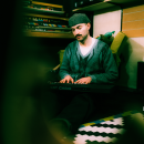 Bandfoto Eyeclimber: Ein Mann sitzt in einem gemütlichen, leicht abgedunkelten Musikstudio auf dem Boden und spielt ein Casio-Keyboard. Er trägt eine dunkle Wollmütze, einen grauen Strickcardigan und ein weißes T-Shirt. Hinter ihm stehen mehrere Keyboards